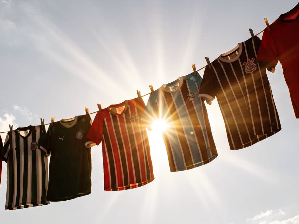 Camisas de clubes de futebol brasileiros penduradas num varal, contra o céu, com o sol brilhando entre elas.