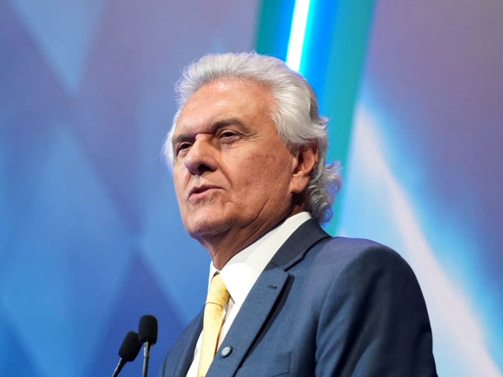 Ronaldo Caiado, de terno azul-marinho, camisa branca e gravata amarela, fala ao microfone diante de um painel azul.